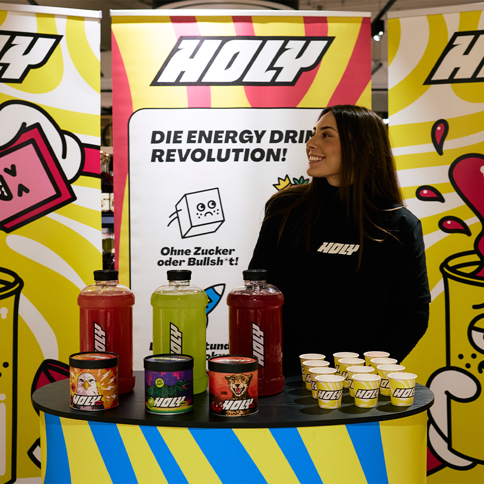 Die Promoterin steht vor dem Holy Energy Stand im Supermarkt und präsentiert die Produktpalette.